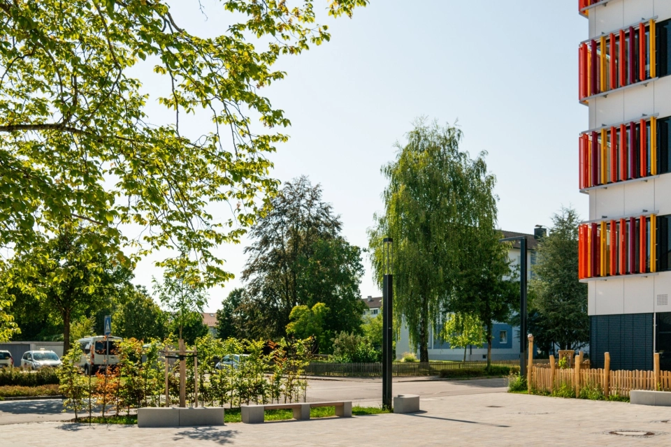 Kalk-Grau und Beige gepflasterter Schulhof mit Sitzelementen und Grünflächen an einem sonnigen Tag mit blauem Himmel. Ein großer Baum spendet Schatten. Im Hintergrund steht ein Schulgebäude mit weißer Fassade und orange-gelben, geometrischen Verzierungen