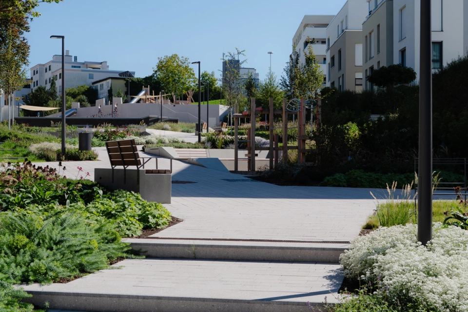 Im Vordergrund befindet sich ein grau gepflasterter Weg mit Stufenelementen und Pflanzbeeten links und rechts davon. Dahinter sind moderne Häuser,  Sitzmöglichkeiten und ein Spielplatz