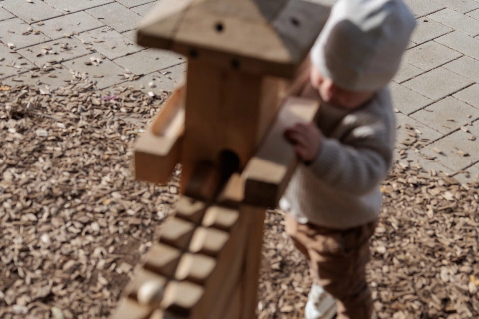 Ein Kind spielt an einer hölzernen Murmelbahn, die auf einem Boden aus Hackschnitzeln steht. Dahinter verläuft ein Weg aus hellen Betonsteinen. 