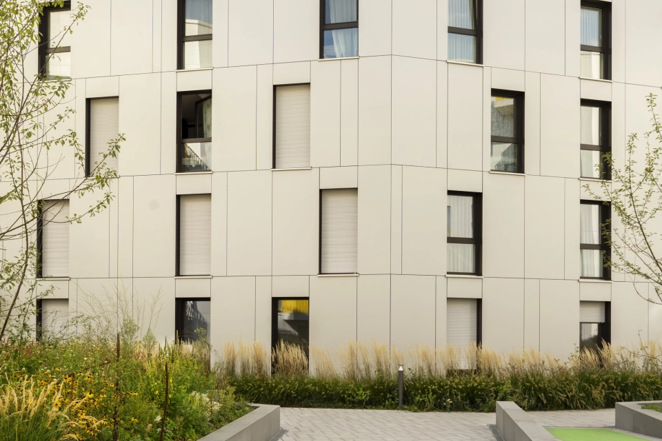 Ein Weg, welcher auf ein Gebäude mit vielen Fenstern und heller Fassade zuführt. Der Weg ist mit basalt- und granitfarbenem GDM.SCENIC individual gepflastert. Außerdem ist er durch Einfassungen begrenzt, welche gleichzeitig als Umrandung für Beete dienen.