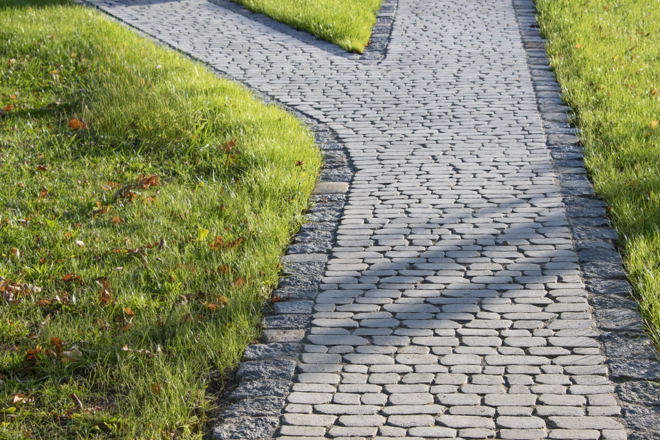 Geschwungener Gartenweg aus sandfarbenem Betonstein, eingebettet in eine gepflegte Grünfläche, führt entlang von Rasen und Bäumen zu einem modernen Holzgebäude.
