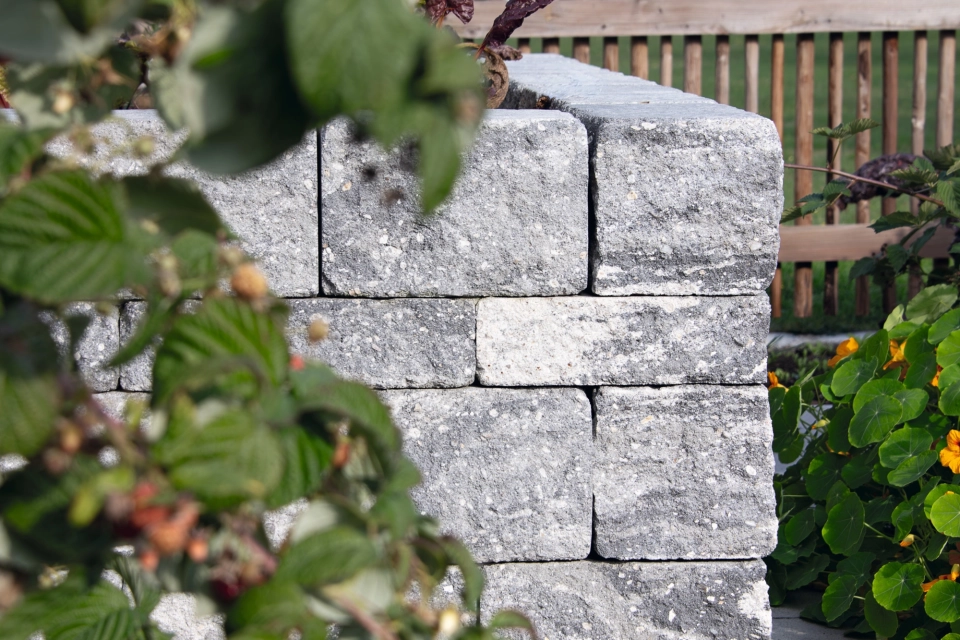 Blick auf eine Ecke eines Hochbeetes. Das Hochbeet ist aus grau-schwarzen Betonsteinen und steht auf gepflastertem Untergrund. Im Hintergrund ist ein Zaun und ein weißes Haus und im Vordergrund sind Äste eines Strauches.