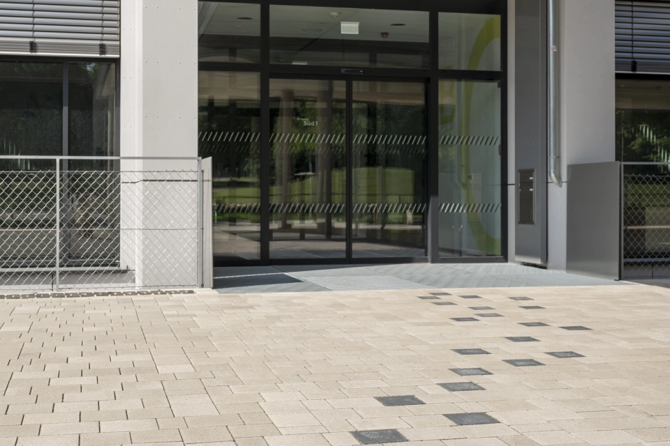 A path consisting of anthracite-colored concrete blocks leads through a beige paved schoolyard to a modern school building. The building has a partly white, partly light gray façade and a large entrance area. 
