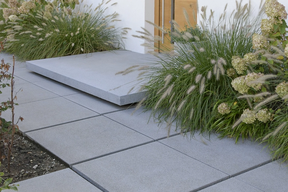 Moderner Hauseingang mit grauem Betonpodest und heller Haustüre aus Holz. Davor sind graue Betonsteinplatten verlegt. Links und Rechts von der Haustüre sind große, grüne Sträucher.