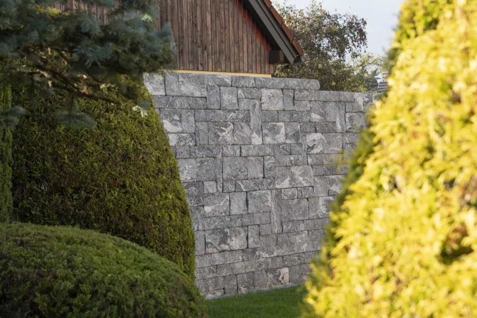 Blick durch grüne und gelbe Bäume und Sträucher hindurch auf die GDM.DECA wall Sichtschutzmauer. Davor befindet sich eine Rasenfläche und im Hintergrund ist ein Holzschuppen.
