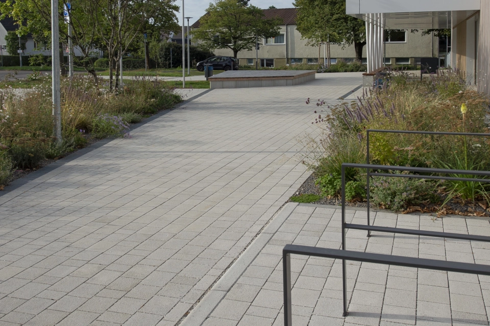 Weg aus Kalk-Grauen, versickerungsfähigen Klimasteinen. Daneben befinden sich anthrazitfarbene Fahrradabstellbügel, Sträucher und Straßenlaternen.
