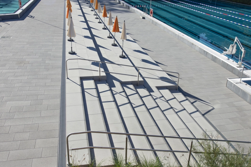 Weitläufiger Bereich eines Freibads mit breiten Sitz- und Zugangstreppen aus GDM.SCENIC step und GDM.SCENIC seat, die entlang des Schwimmbeckens verlaufen; daneben stehen geschlossene Sonnenschirme in einer Reihe.