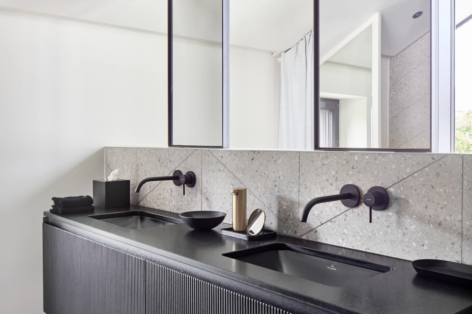 in modernes Badezimmer mit schlichtem Interieur, ausgestattet mit zwei Waschbecken und einem schwarzen/anthrazitfarbenen Unterschrank. Der Boden und die Wandfliesen in Fischgrät-Optik aus Terrazzo-Beton verleihen dem Raum eine elegante Note.