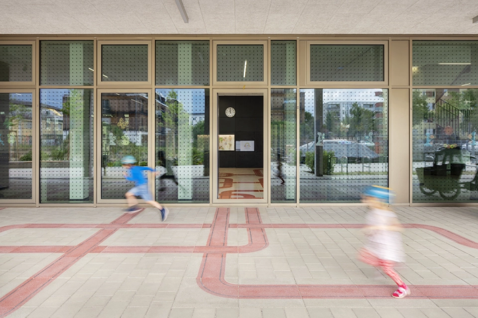 Eingangsbereich eines Schulgebäudes mit großen Fenstern. Davor sind zwei Kinder zu erkennen, welche auf einer grau-beigen Pflasterfläche herum laufen. Durch diese zieht sich ein Band aus roten Betonsteinen.