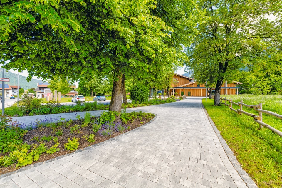 Modern gepflasterter Weg mit großer Grünfläche und Bäumen vor einem Bürgerzentrum  mit Holzfassade