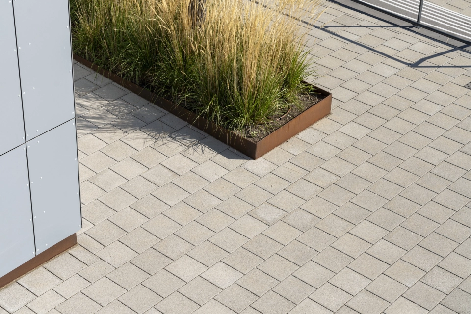 Nahaufnahme der gepflasterten Fläche aus Betonstein. Daneben ist ein Beet mit Gräsern und einer Einfassung aus Cortenstahl. 