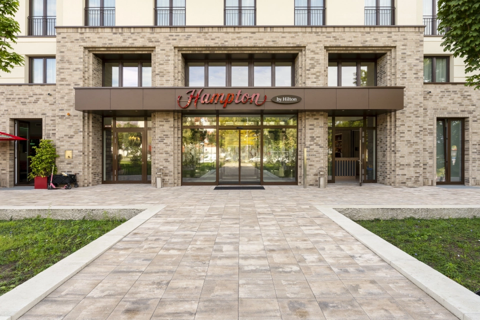 Moderne Gebäudefassade mit braunen Wandfliesen, vielen großen Fenstern und dunkelbraunem Schild über dem Eingang. Davor ist ein gepflasterter Weg aus muschelkalkfarbenen Pflastersteinen - dem GDM.KLIMASTEIN. Daneben befinden sich Grünflächen.