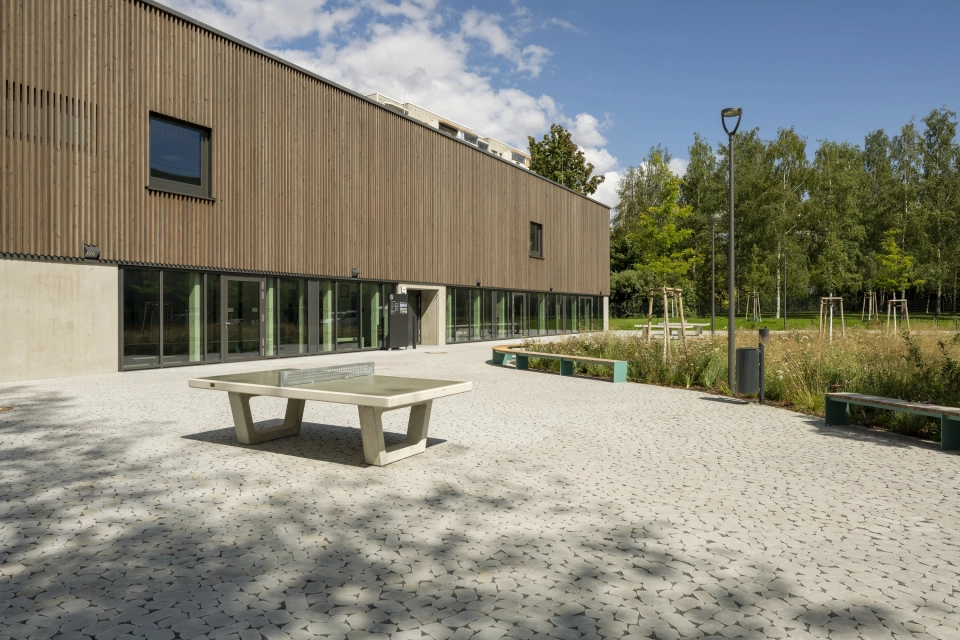 Ein mit rundlich und natürlich wirkenden Betonsteinen gepflasterter Schulhof, welcher rechts neben einem modernen Schulgebäude mit Holz-Fassade liegt. Auf dem Platz ist eine Tischtennisplatte und im Hintergrund eine bewachsene Wiese mit Streuchern zu erkennen.