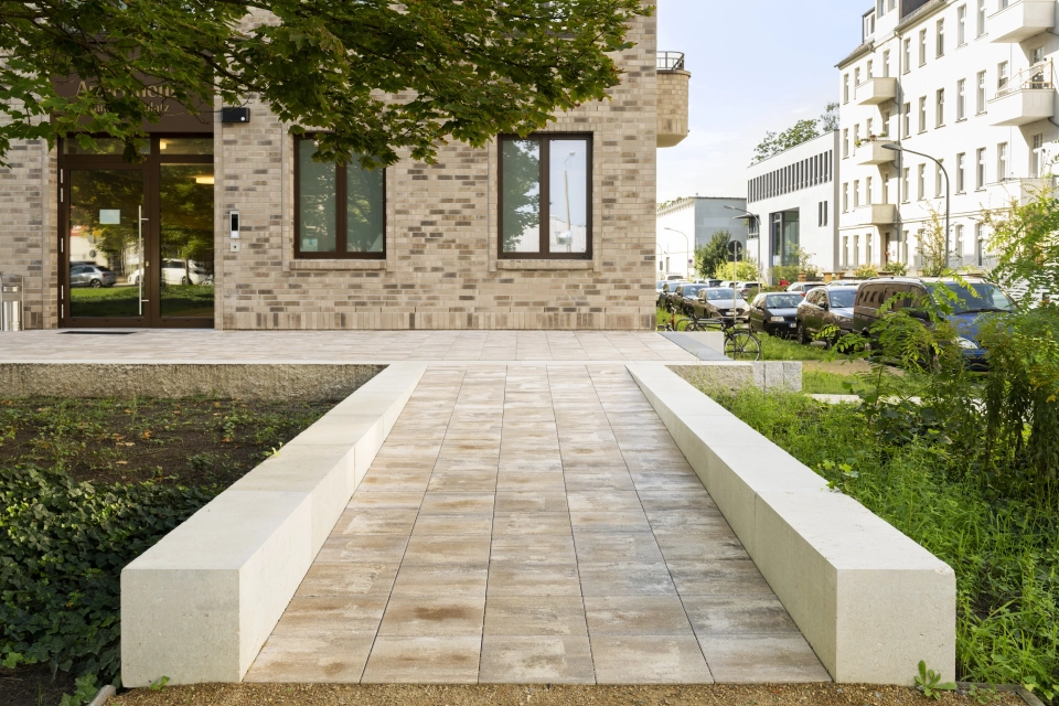 Blick auf einen gepflasterten, kalksteinfarbenen Gehweg mit GDM.MAXI block als Wegbegrenzung. Der Weg führt auf ein Gebäude mit braunen Wandfliesen zu. Drumherum sind grüne Bäume und Sträucher zu finden.