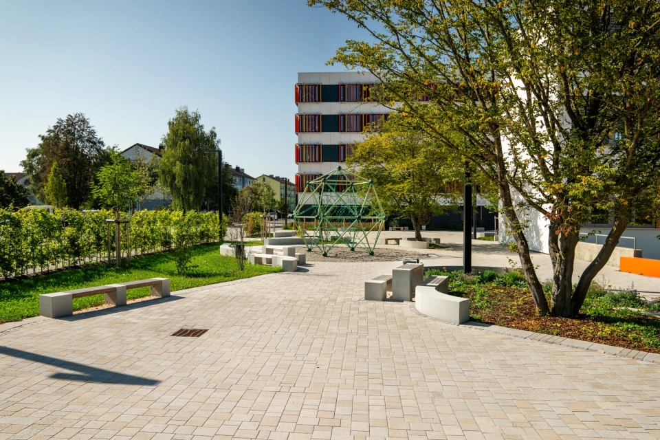 Kalk-Grau und Beige gepflasterter Schulhof mit vielen Sitzelementen und Grünflächen an einem sonnigen Tag mit blauem Himmel. Ein Klettergerüst innerhalb einer Sandfläche ziehrt den Hof und große Bäume spenden Schatten. Im Hintergrund steht ein Schulgebäude mit weißer Fassade und orange-gelben, geometrischen Verzierungen