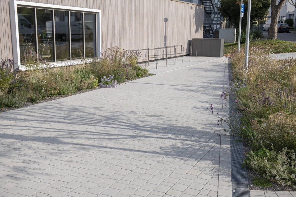 Weg aus Kalk-Grauen, versickerungsfähigen Klimasteinen. Daneben befinden sich Sträucher und Gräser und im Hintergrund findet man anthrazitfarbene Fahrradabstellbügel.