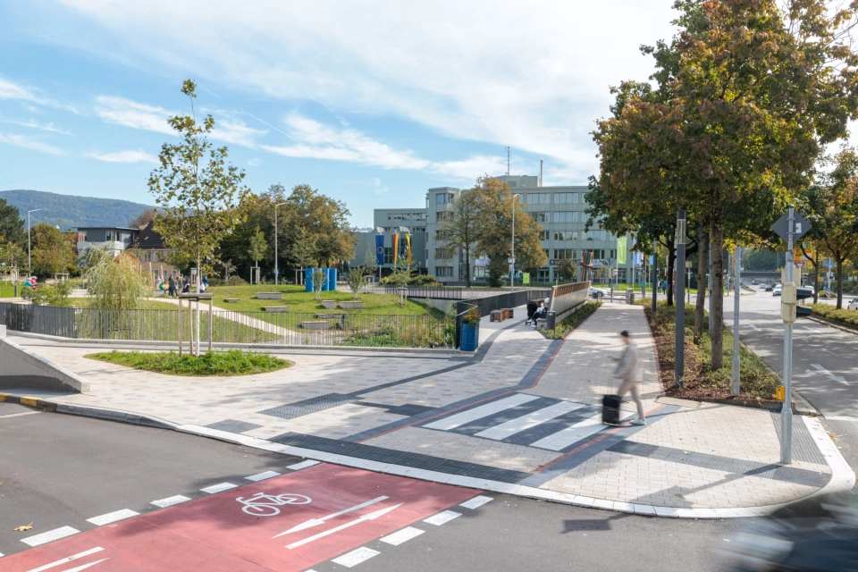 Übersicht einer städtischen Weganlage, in der ein breiter Gehweg überwiegend mit beigem GDM.KLIMASTEIN gepflastert ist. Zur Markierung des Querungsbereichs sind rote und weiße Pflastersteine aus GDM.TETRAGO stone integriert. Die Wege grenzen an eine begrünte Parkfläche mit Rasen, Bäumen, Sitzbereichen und einem Wasserlauf. Im Vordergrund verläuft ein Radweg.