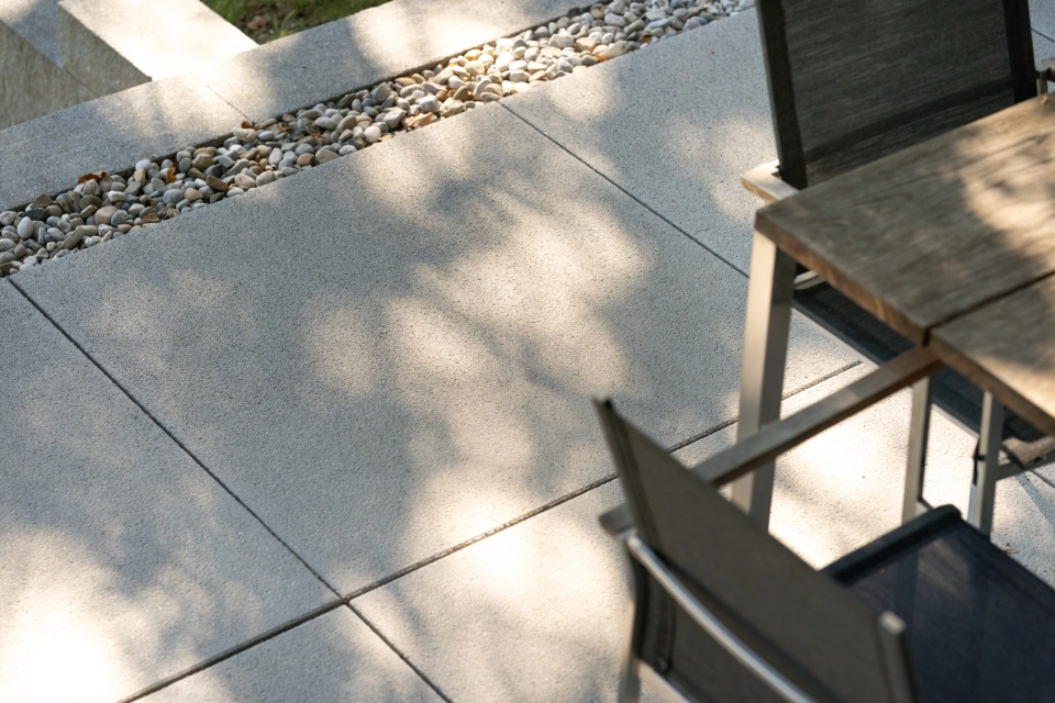 Nahaufnahme von grauen GDM.NUEVA light Terrassenplatten neben einer Kiesspur sowie Teilen eines Outdoor‑Tisches und Stühlen, mit sanften Schatten von umliegender Bepflanzung.