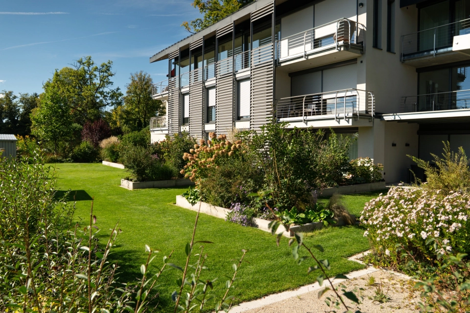 Außenaufnahme einer modernen Wohnanlage mit mehreren Balkonen und großen Fensterfronten. Davor liegt eine weitläufige, gepflegte Grünfläche mit dicht bepflanzten Beeten aus verschiedenen Sträuchern, Ziergräsern und blühenden Pflanzen. Im Hintergrund stehen weitere Bäume unter klarem blauem Himmel.