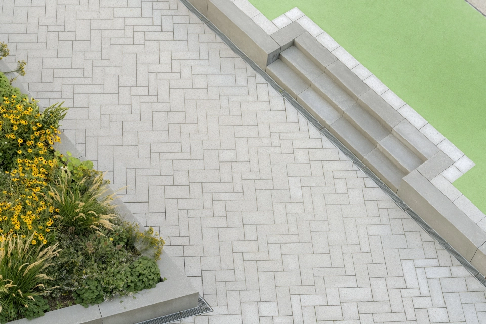 Mit basalt- und granitfarbenem GDM.SCENIC individual gepflasterter Weg. Am rechten Rand ist ein bepflanztes Beet und links ist eine kleine Treppe, welche auf eine erhöhte Fläche mit grünem Bodenbelag führt. 