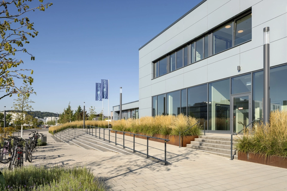 Blick auf ein graues Gebäude mit großer Fensterfront. Auf dem Vorplatz ist der GDM.SCENIC individual verlegt. Auch finden sich Treppen und Beete mit Gräsern.
