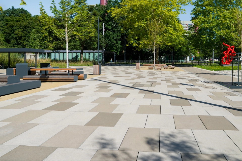 Offener Platz auf einem Universitätscampus mit Bäumen, Sitzbänken und einer roten Skulptur im Hintergrund, wobei der Platz mit GDM.SCENIC stone in drei unterschiedlichen Farbabstufungen gepflastert ist.