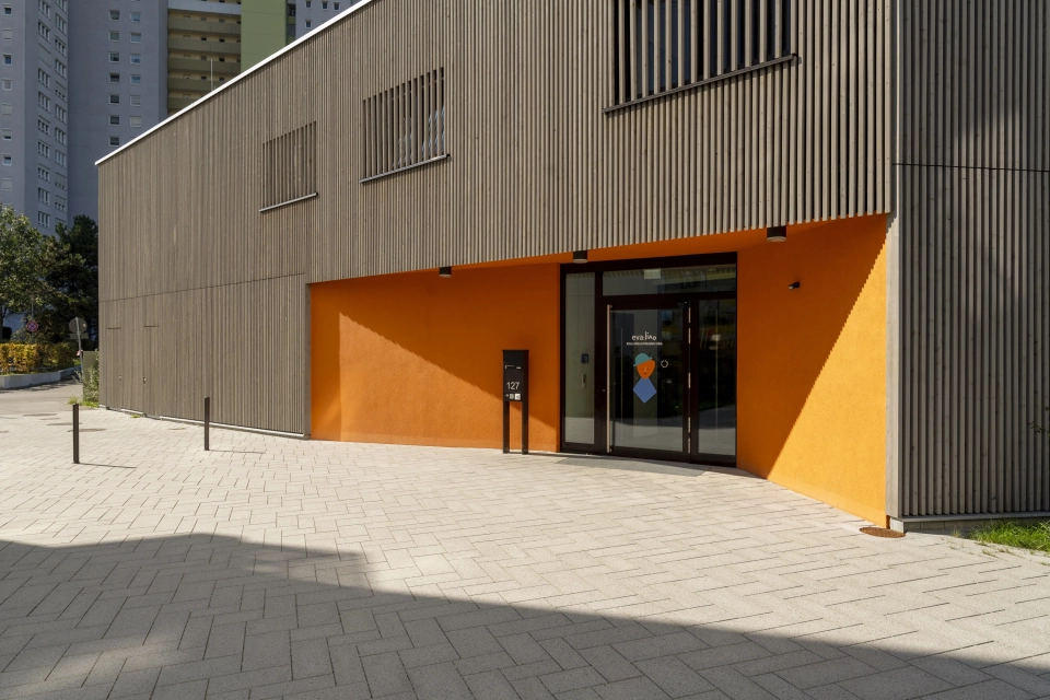 Großes Gebäude mit Holzverkleidung. Der Eingangsbereich hat einen orangenen Anstrich. Der Vorplatz ist mit basalt- und granitfarbenem GDM.SCENIC individual gepflastert.