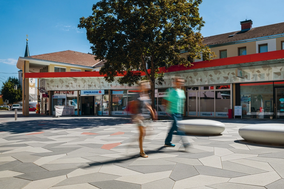 Vorplatz mit Baum, rund geschwungenen Sitzelementen und Belag aus mehrfarbigen Polygonalplatten vor Einkaufszentrum
