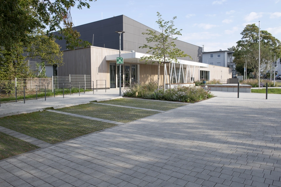 Moderne Außenfassade einer Sporthalle mit anthrazitfarbenen Platten im oberen Bereich und Holzverkleidung im unteren Bereich. Davor befindet sich ein mit Kalk-Grauen, versickerungsfähigen Betonsteinen gepflasterter Vorplatz mit Parkplätzen aus Rasengittersteinen und Sträuchern, Gräsern und Bäumen. 