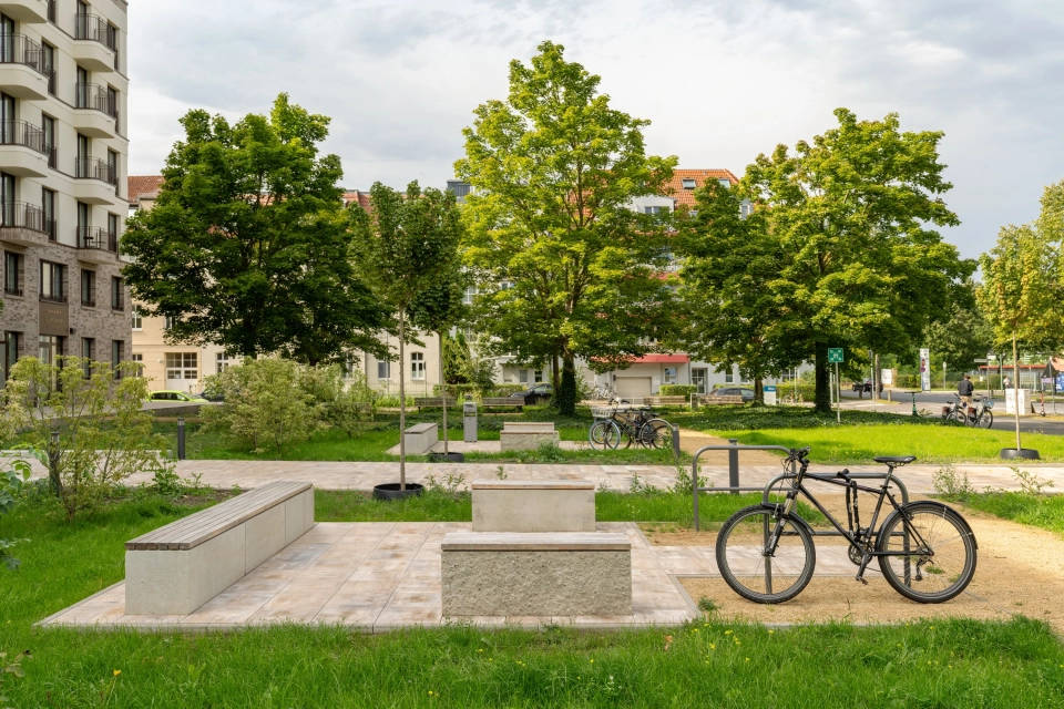 Grüne Landschaft mit Bäumen und Häusern im Hintergrund. Im Vordergrund ist eine gepflasterte Fläche mit Sitzblöcken aus Beton und Holzsitzfläche. Daneben befinden sich Fahrradständer mit einem Fahrrad.