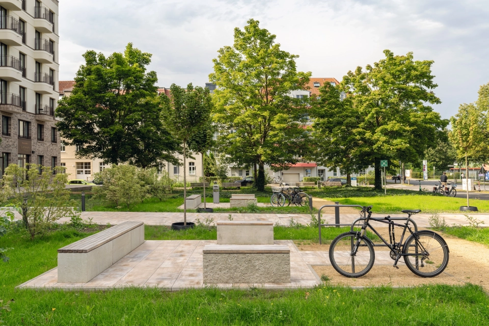 Grüne Landschaft mit Bäumen und Häusern im Hintergrund. Im Vordergrund ist eine gepflasterte Fläche mit Sitzblöcken aus Beton und Holzsitzfläche. Daneben befinden sich Fahrradständer mit einem Fahrrad.