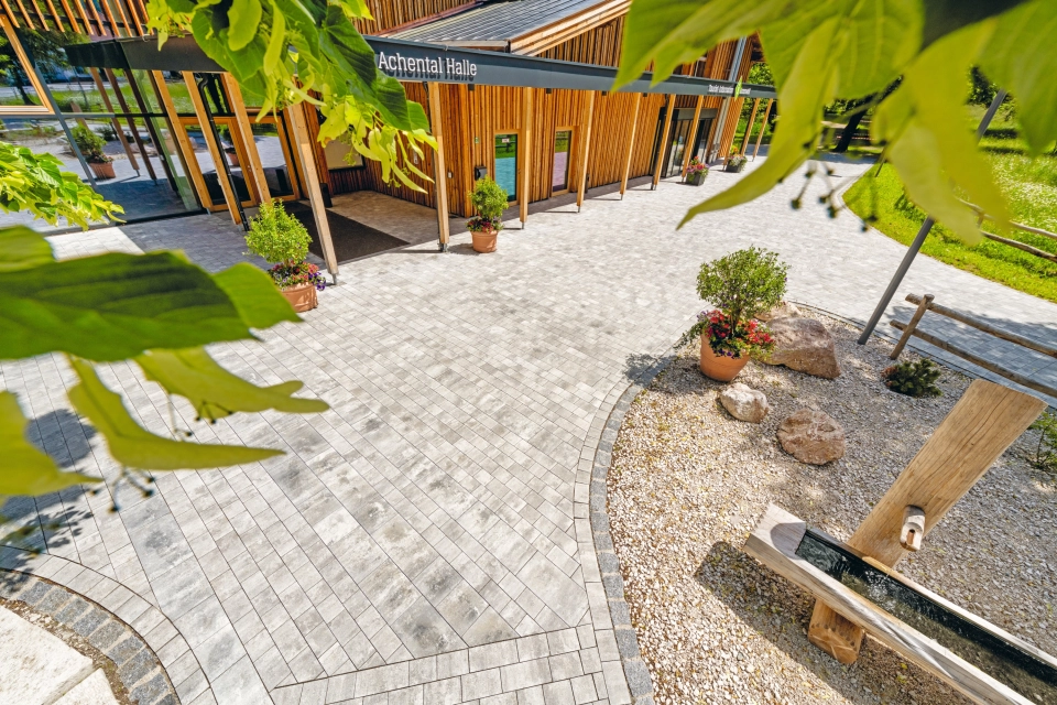 Modern gepflasterter Platz mit großer Grünfläche, Bäumen und Sitzelementen vor einem Bürgerzentrum  mit Holzfassade