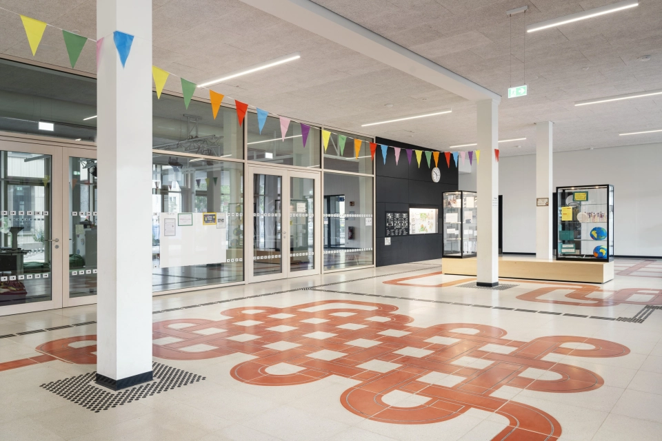 Der Eingangsbereich eines Schulgebäudes, durch dessen grau-beigen Schulboden sich ein rotes Band aus Sichtbeton-Platten zieht.