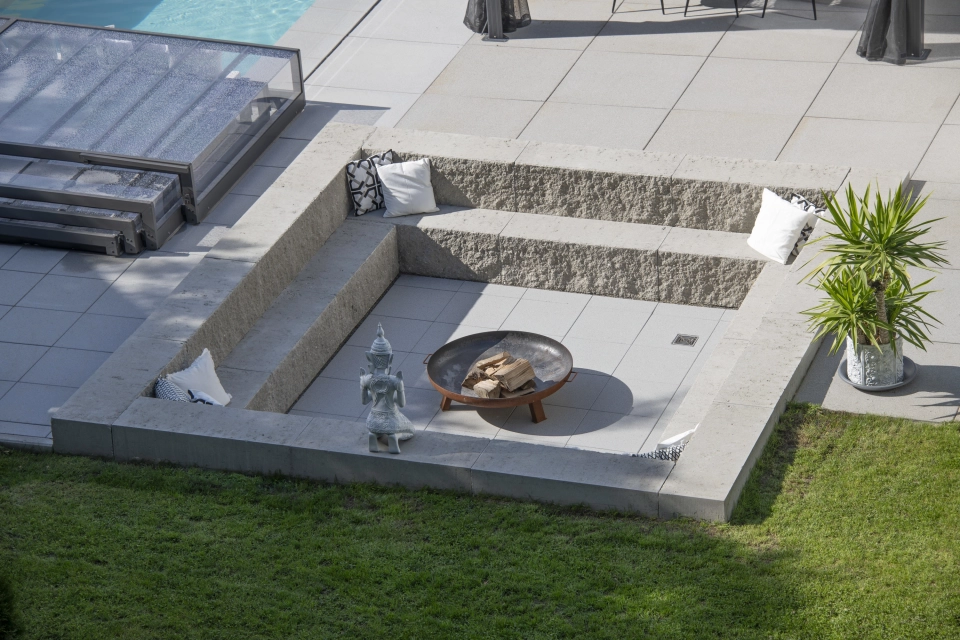 Quadratische, zweistufige Sitzmöglichkeit aus GDM.MAXI block mit einer Feuerschale in der Mitte. Daneben ist eine Rasenfläche und eine Terrasse mit GDM.NUEVA light. Auf der Terrasse ist eine zusammengeklappte Poolabdeckung.