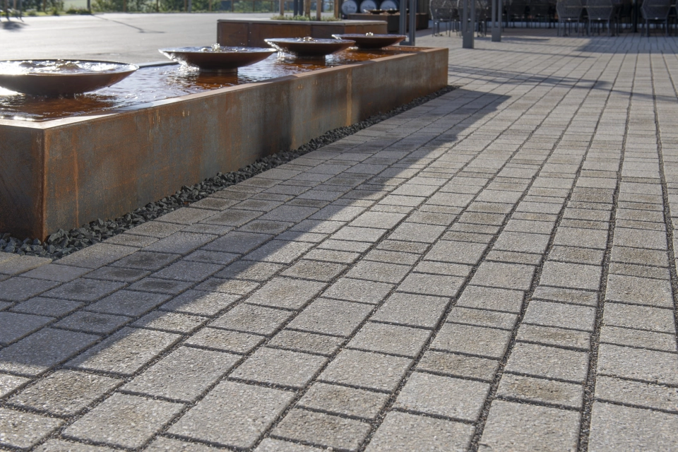 Nahaufnahme von Naturbeige schattierten Pflastersteinen mit schiefer Fuge. Daneben befindet sich ein Wasserbecken mit Schalen aus rostigem Corthenstahl.