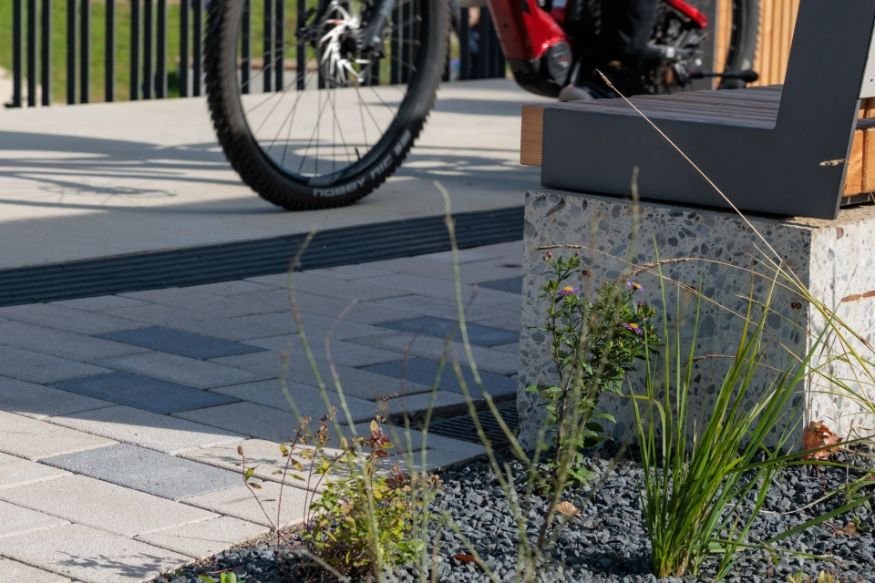 Close-up view of a planted area with ornamental grasses and dark chippings in the foreground. Behind it is a path made of beige GDM.KLIMASTEIN with individual grey paving stones scattered as accents. On the right is a modern bench with a concrete base; a bicycle can be seen in the background.