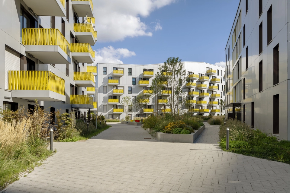 Großer Innenhof mit grün bewachsenen Beeten. Dieser ist mit dem basalt- und granitfarbenem GDM.SCENIC individual gepflastert. Ringsherum sind große Wohngebäude mit heller Fassade und gelben Balkonbrüstungen. 