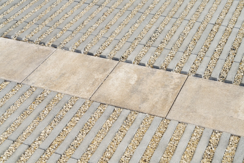 Nahaufnahme von schräg verlaufenden, grauen Fugensteinen, dem GDM.SCADA liner. Zwischen den einzelnen Steinen ist Kies zu finden. Zusätzlich ist der muschelkalkfarbene GDM.KLIMASTEIN als Abgrenzung verlegt.