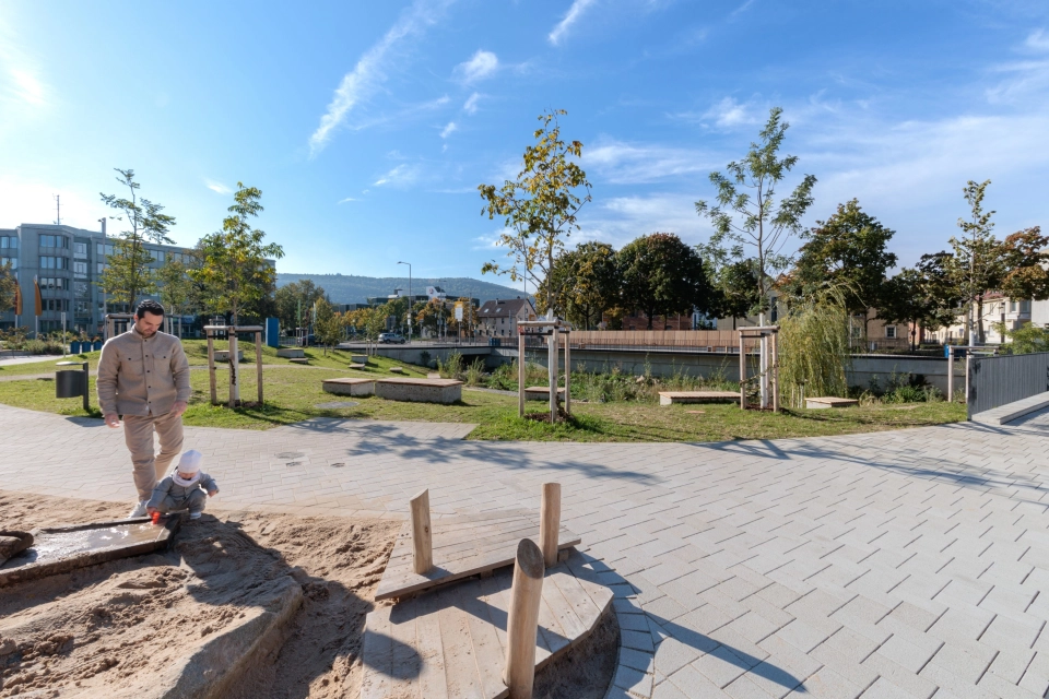 Offen gestalteter Außenbereich mit einem Weg aus Betonsteinen, der an einen Sandspielbereich mit Holzelementen grenzt. Im Vordergrund befindet sich eine erwachsene Person mit einem kleinen Kind, im Hintergrund liegen Grünflächen, junge Bäume und ein Wasserlauf mit Uferbepflanzung.