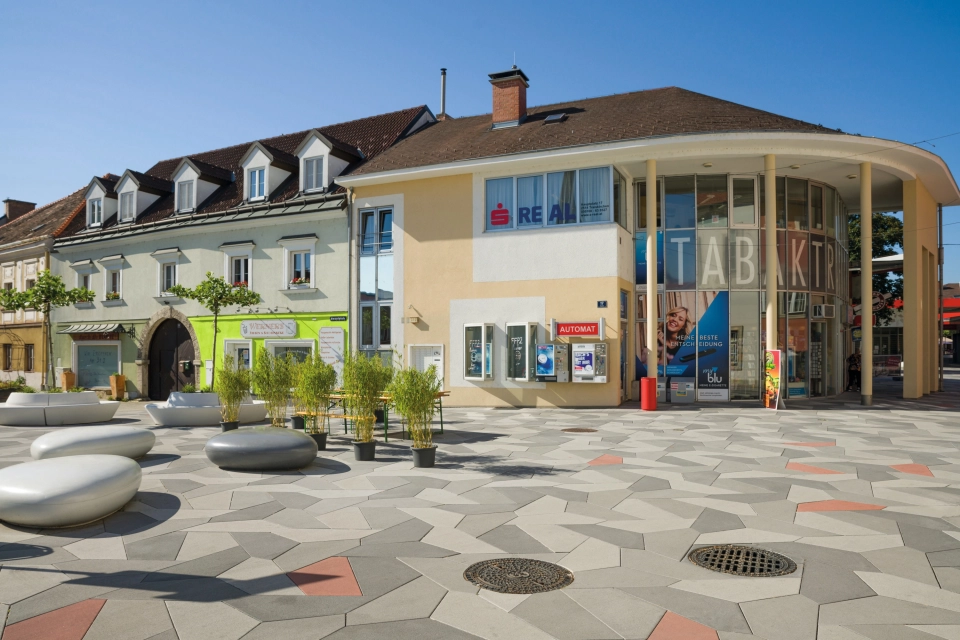 Vorplatz mit Begrünung, rund geschwungenen Sitzelementen und Belag aus mehrfarbigen Polygonalplatten