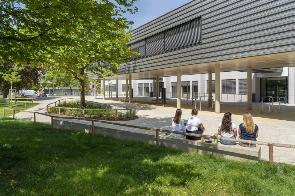 The schoolyard is surrounded by green meadows, trees and seating elements where children can chat with each other. A modern, multi-part school building can be seen in the background. The front part stands on concrete pillars and is clad in dark gray. The rear part has a white façade. 