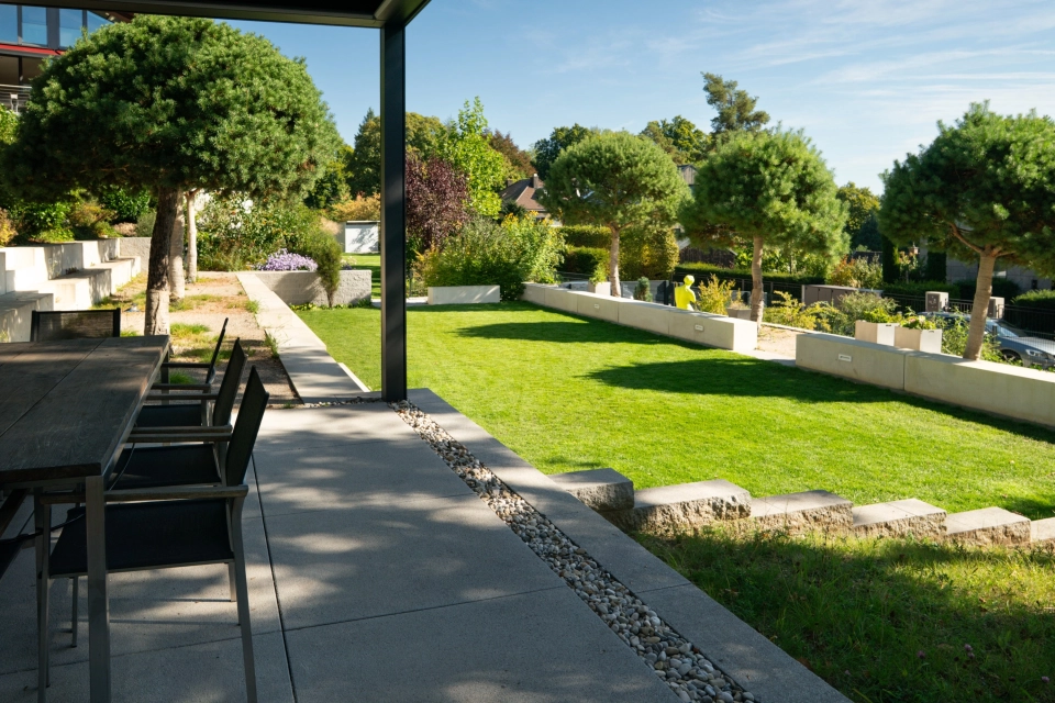Moderne Gartenterrasse mit überdachtem Sitzbereich und hellen GDM.NUEVA light Terrassenplatten, angrenzend an eine gepflegte Rasenfläche, umgeben von niedrigen Mauern, Zierkies und formgeschnittenen Bäumen.