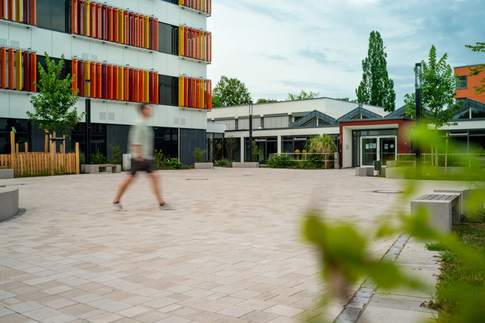Ein Mann geht über einen ansprechenden Schulhof mit vielen Sitzelementen und Grünflächen. im Vordergrund sind unscharf ein paar Zweige mit grünen Blättern zu erkennen. Im Hintergrund steht ein Schulgebäude mit weißen Fassaden und orange-gelben, geometrischen Verzierungen