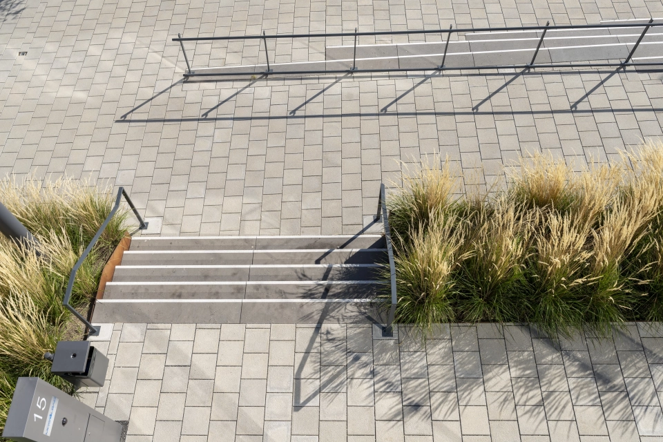 Blick von oben auf eine gepflasterte Fläche aus grauem GDM.SCENIC individual und eine anthrazitfarbene Treppe. Neben der Treppe sind Beete mit Ziergras.