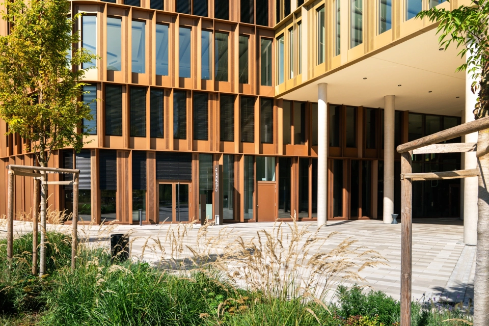 Großes Bürogebäude mit Fensterfassade. Auf dem Vorplatz ist kalksteinfarbener GDM.SCENIC individual verlegt. Im Vordergrund ist ein Beet mit grünen Pflanzen und kleinen Bäumen.