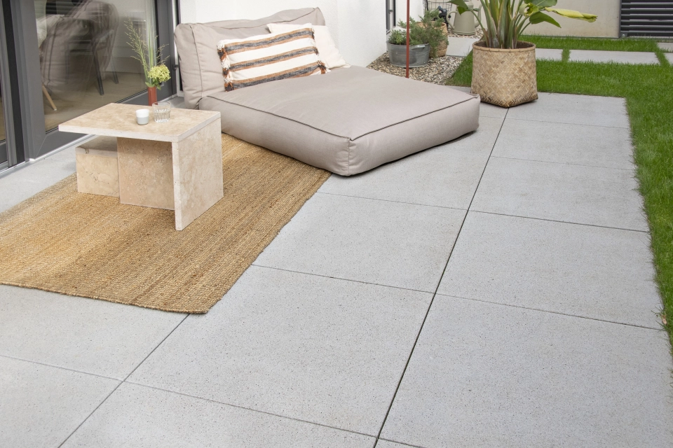 Nahaufnahme einer Terrasse mit grauen Terrassenplatten aus Beton. Darauf ist ein beiges Liegekissen, ein brauner Teppich und ein Tisch. 