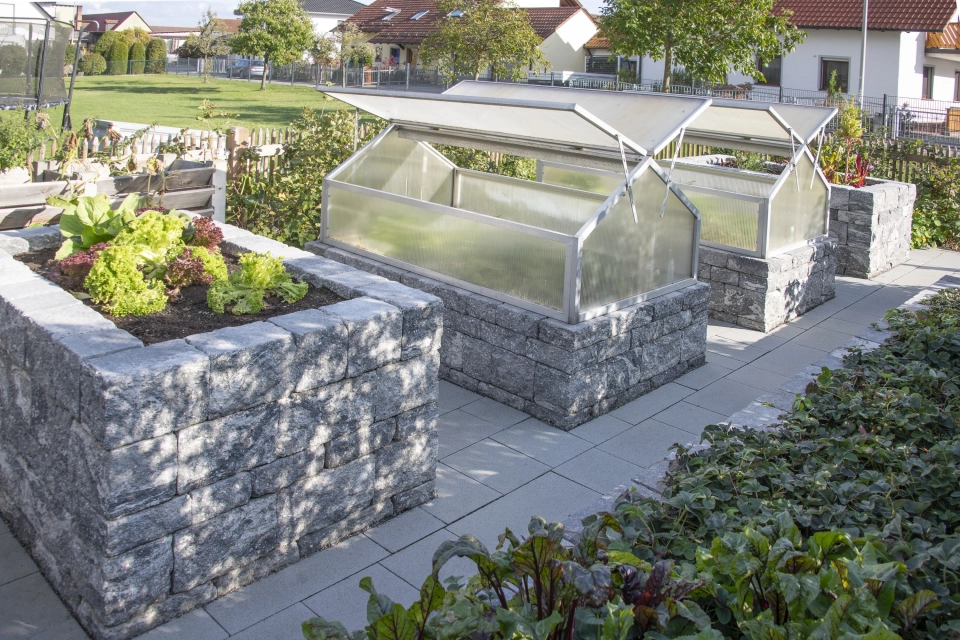4 klassische Hochbeete aus GDM.DECA wall auf gepflastertem Untergrund. Im vordersten Beete wächst Salat. Die beiden mittleren Hochbeete haben ein integriertes Gewächshaus. Im Hintergrund sind Bäume und Häuser zu erkennen und im Vordergrund sind grüne Sträucher.