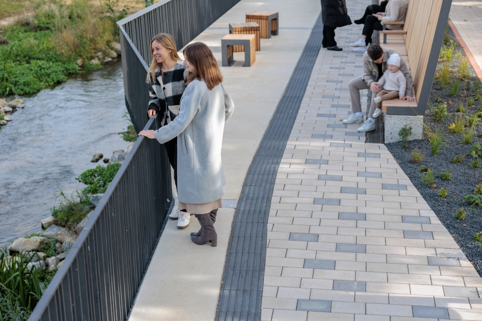Promenade entlang eines Bachlaufs mit Sitzgelegenheiten und einem Weg aus hellem GDM.KLIMASTEIn, in den regelmäßig graue Pflastersteine integriert sind. Entlang des Geländers stehen mehrere Personen, weitere Personen sitzen auf den Holzsitzbänken im Hintergrund.