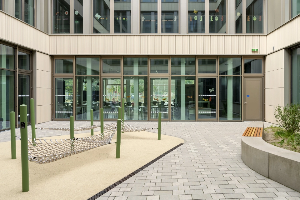 Innenhof eines Schulgebäudes mit großen Fenstern, ausgestattet mit kleinen Klettergeräten und Sitzelementen. Gepflastert ist er mit grau-beigen, versickerungsfähigen Betonsteinen.