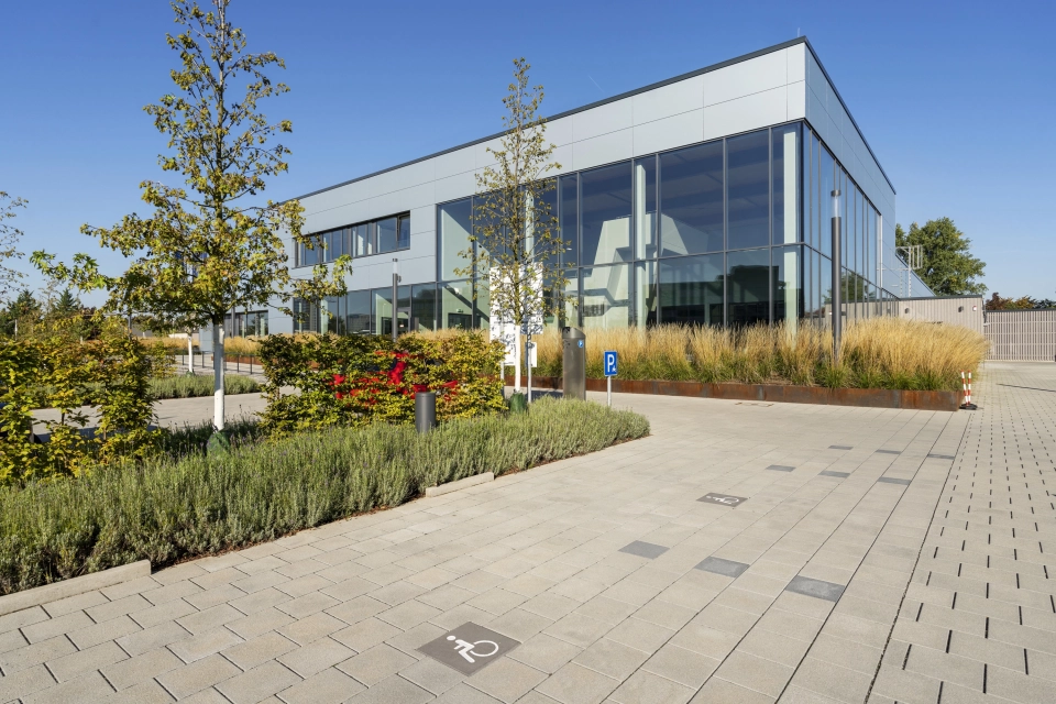 Im Vordergrund ist ein grau, gepflasterter Parkplatz. Daneben befinden sich mehrere Beete mit verschiedenen Bäumen, Gräsern und Sträuchern. Im Hintergrund ist ein großes Gebäude mit grauer Fassade und großer Fensterfront.